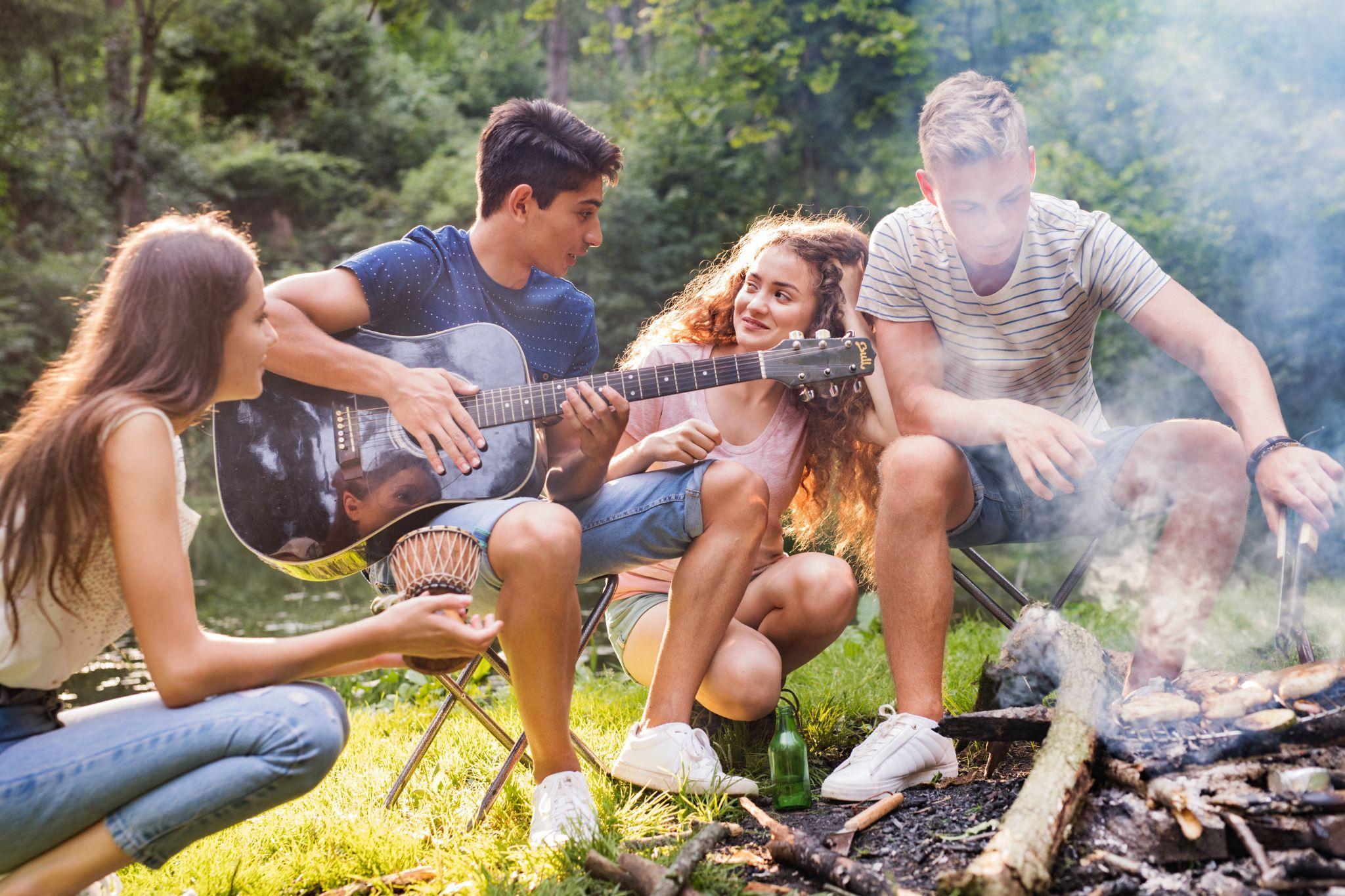 Group of teenagers camping in forest, sitting by the open fire, cooking and playing guitar.