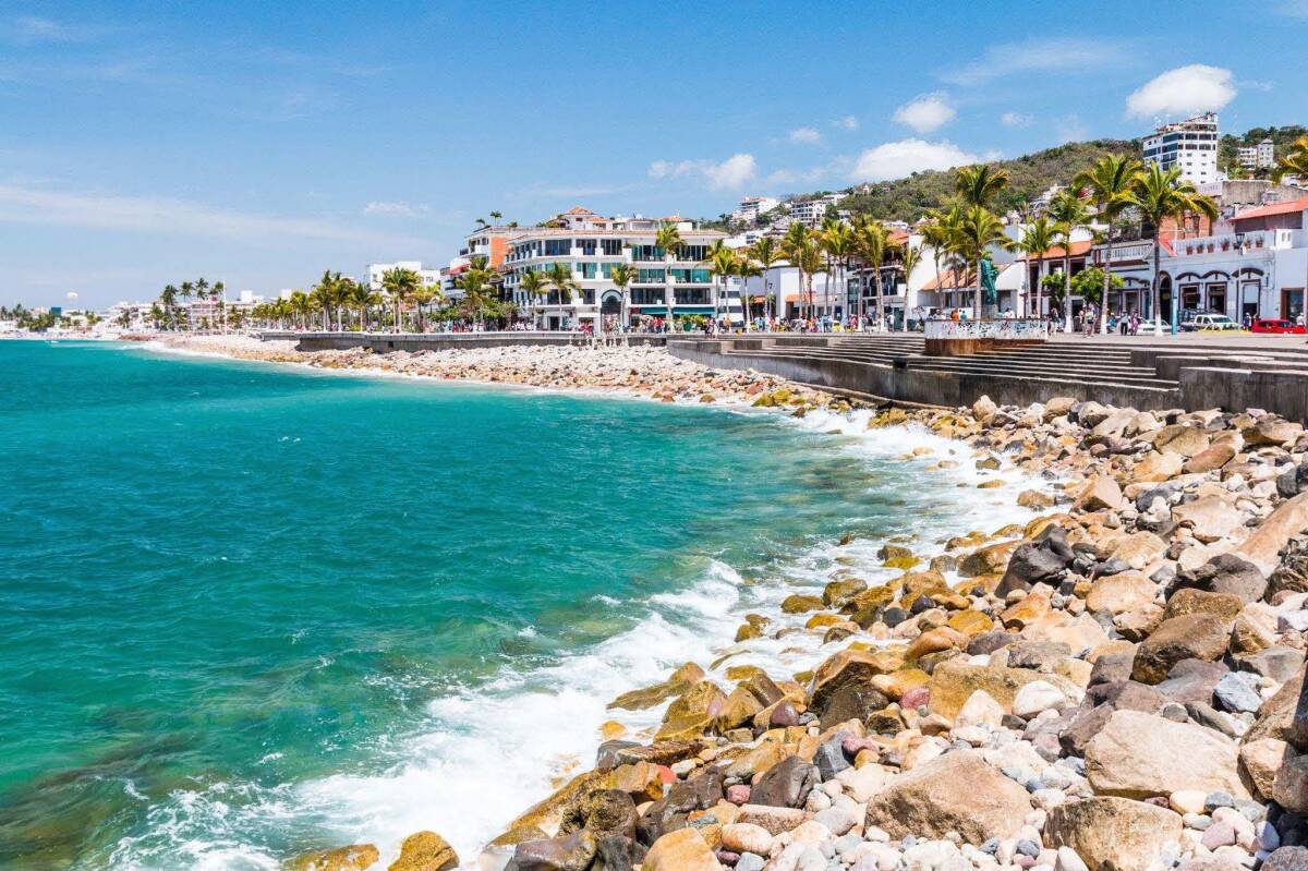 A sunny day in Puerto Vallarta, Mexico, highlighting the rocky shoreline, vibrant blue waters, and a bustling waterfront with palm trees and modern buildings.