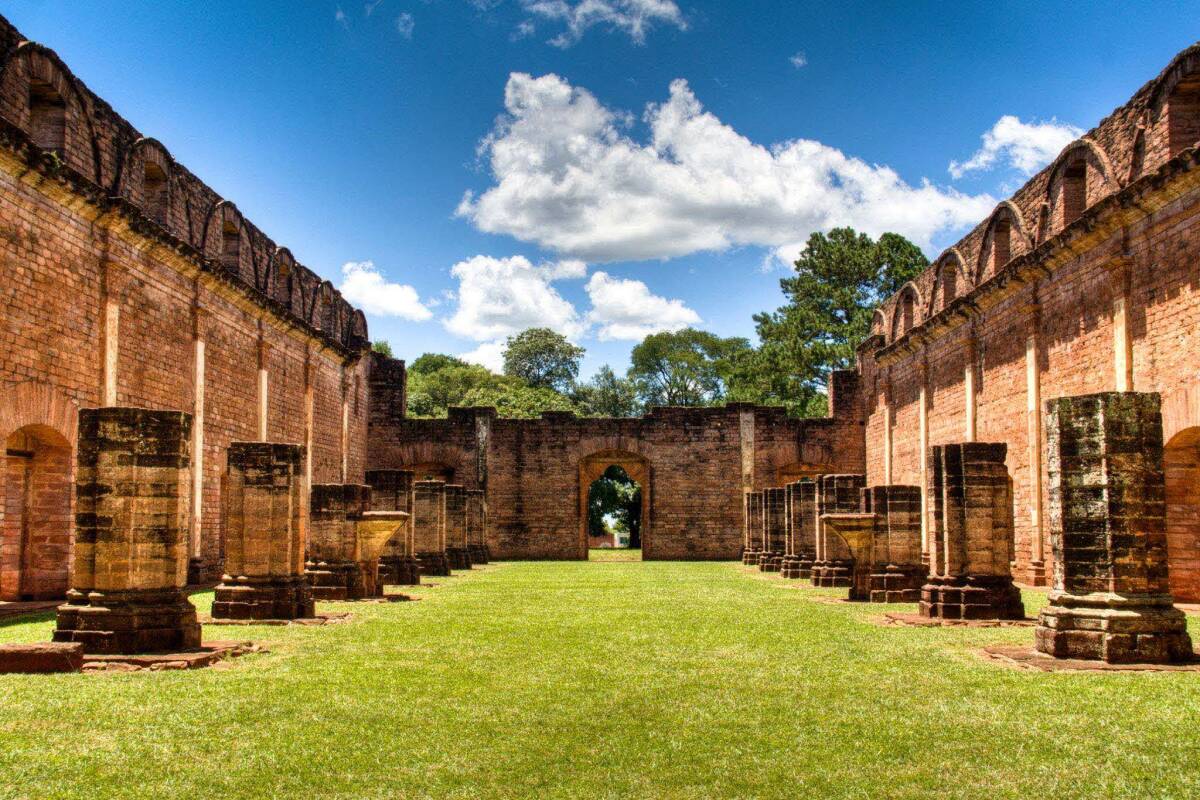 Ancient Jesuit ruins in Paraguay with weathered stone columns and walls surrounded by lush green grass under a bright blue sky.